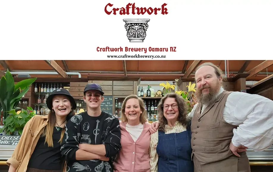 Craftwork Brewery staff smiling behind bar in Oamaru, NZ with bottles, plants, and warm rustic decor in the background.
