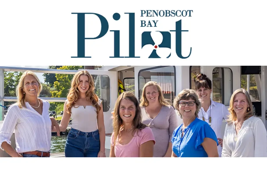 A group of smiling women stands in front of a boat under the Penobscot Bay Pilot logo, representing the PenBay Pilot team.