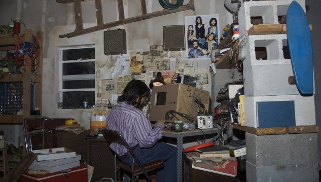 A recreated 1970s garage workshop with a man working at a wooden-cased computer, surrounded by electronics, shelves, magazine clippings and a band poster.